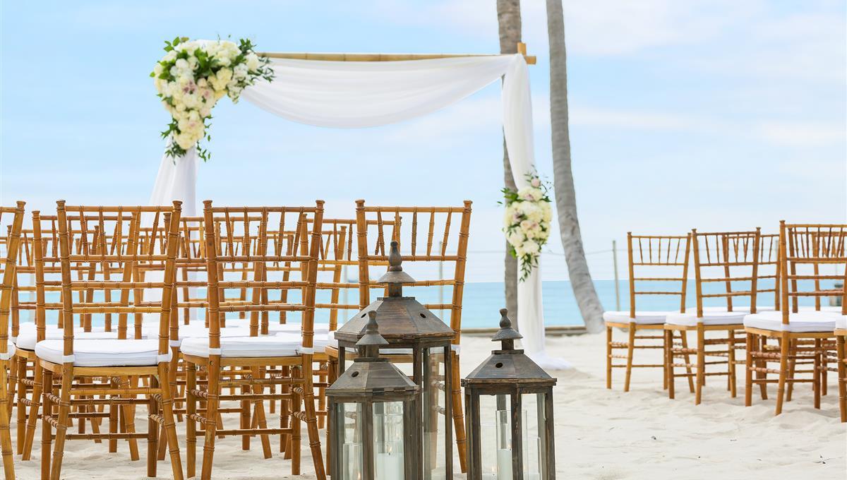Wooden chairs are arranged on sandy beach with a floral-decorated arch nearby; lanterns hold candles in front, with palm trees and ocean visible in Key West, The Keys.